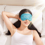 A woman in a white tank top smiles while lying on a white bed, wearing a light blue sleep mask with a white logo.