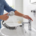 A person's hands operating the chrome shower spray head, attached to a sink faucet, with water flowing into the basin.