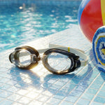Black and clear swimming goggles with a beige strap on blue pool tiles next to a beach ball and towel.