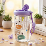 A clear glass tea sipper cup with a purple lid and cartoon duck face design, filled with water, sits on a wooden table with