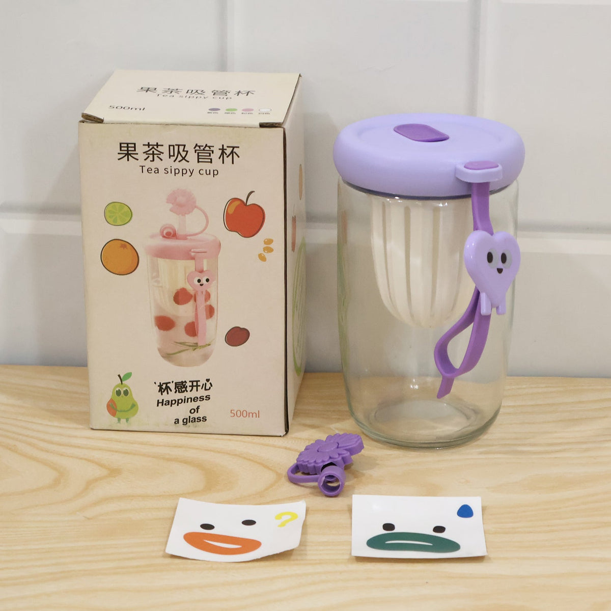 A glass tea sipper cup with a purple lid, a box, and two cartoon face stickers are displayed on a wooden surface.