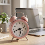 Pink twin bell alarm clock on a wooden desk, with a laptop and a small green plant in soft lighting.