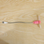 Pink USB mini fan with white blades and a flexible silver gooseneck, laid flat on a light wooden surface.