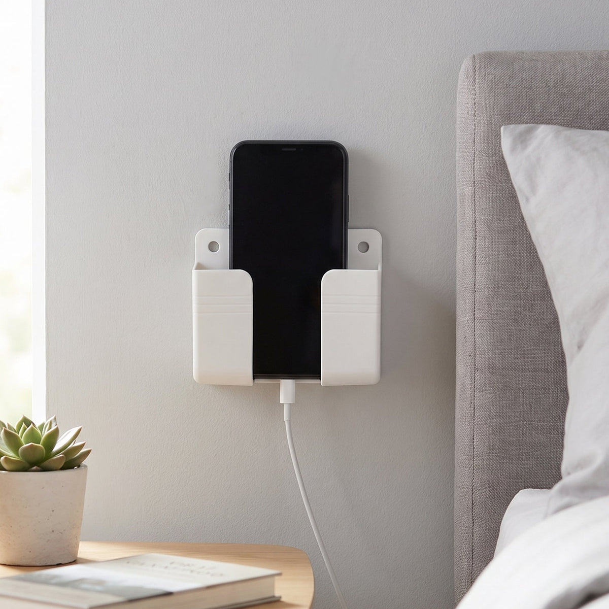 A white wall-mounted phone holder with a black smartphone charging next to a bed and nightstand.