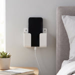 A white wall-mounted phone holder with a black smartphone charging next to a bed and nightstand.