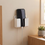 A white wall-mounted phone holder with a black smartphone charging, positioned on a light brown wall near a window.