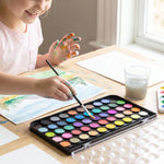 A child's hands, stained with paint, using a brush to paint with a 48-color watercolor set on a wooden table.