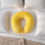 A bright yellow U-shaped neck pillow with snap buttons lies on a white bedspread with pillows and a beige blanket.