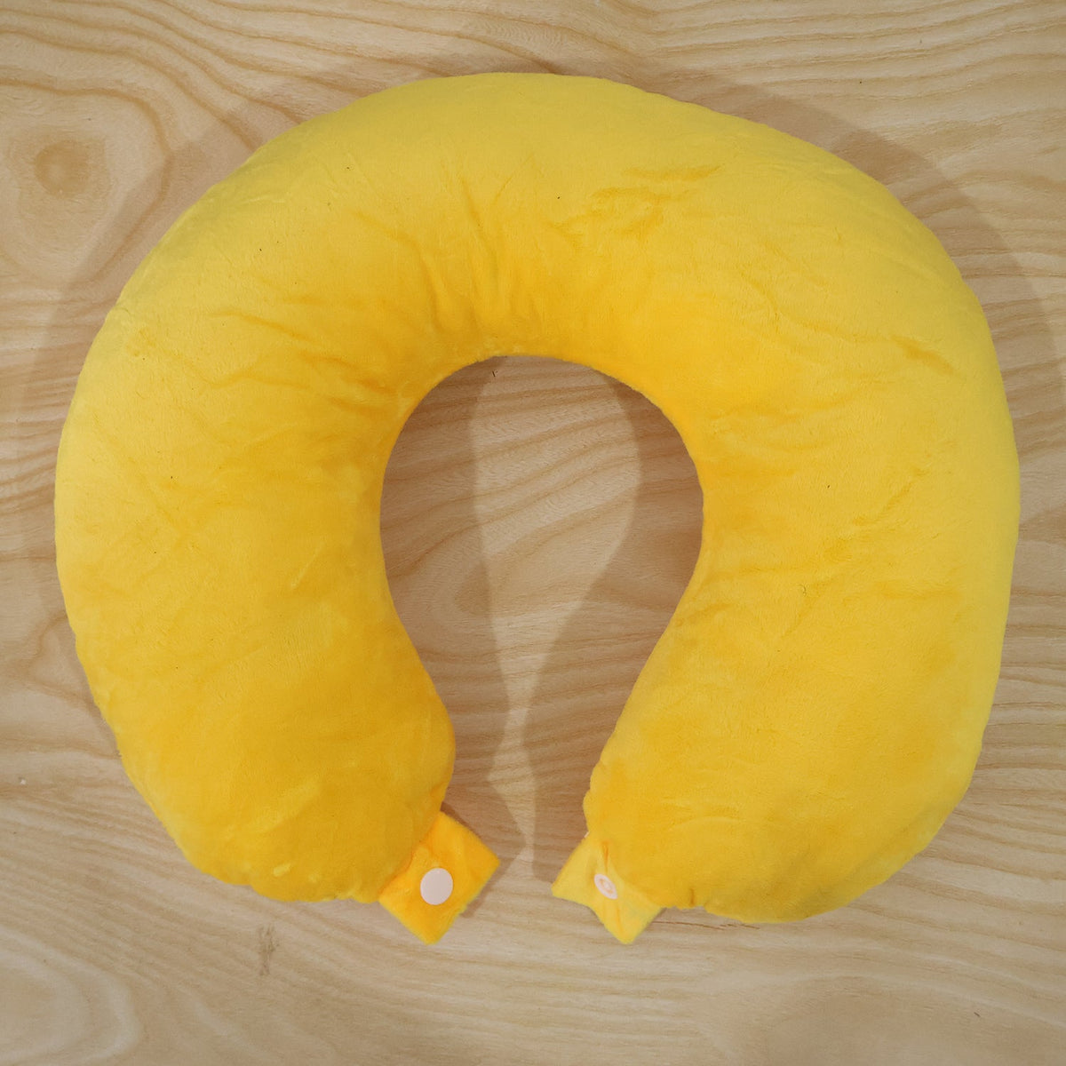 A bright yellow U-shaped neck pillow with two white snap buttons is laid flat on a light brown wooden surface.