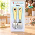 A yellow and silver panda-themed spoon and fork set is packaged in clear plastic on a wooden table.