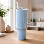 A light blue travel tumbler with a handle and straw sits on a wooden table next to a white vase with flowers.