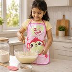 A young girl in a pink monkey apron smiles while whisking batter in a glass bowl in a kitchen.
