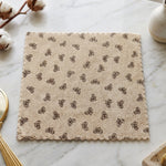 A beige microfiber towel with a dark bow pattern and scalloped edges lies flat on a white marble surface, with cotton