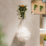 A green cartoon wolf hook holds a white bath pouf on a tiled bathroom wall, with an inset of two wolf hooks on a desk.