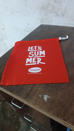 A red drawstring bag with "LET'S SUMMER" and Havaianas logo, resting on a dark wooden desk with metal handles.