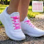 A person wearing white sneakers with bright pink silicone no-tie shoelaces, walking on a gravel path.