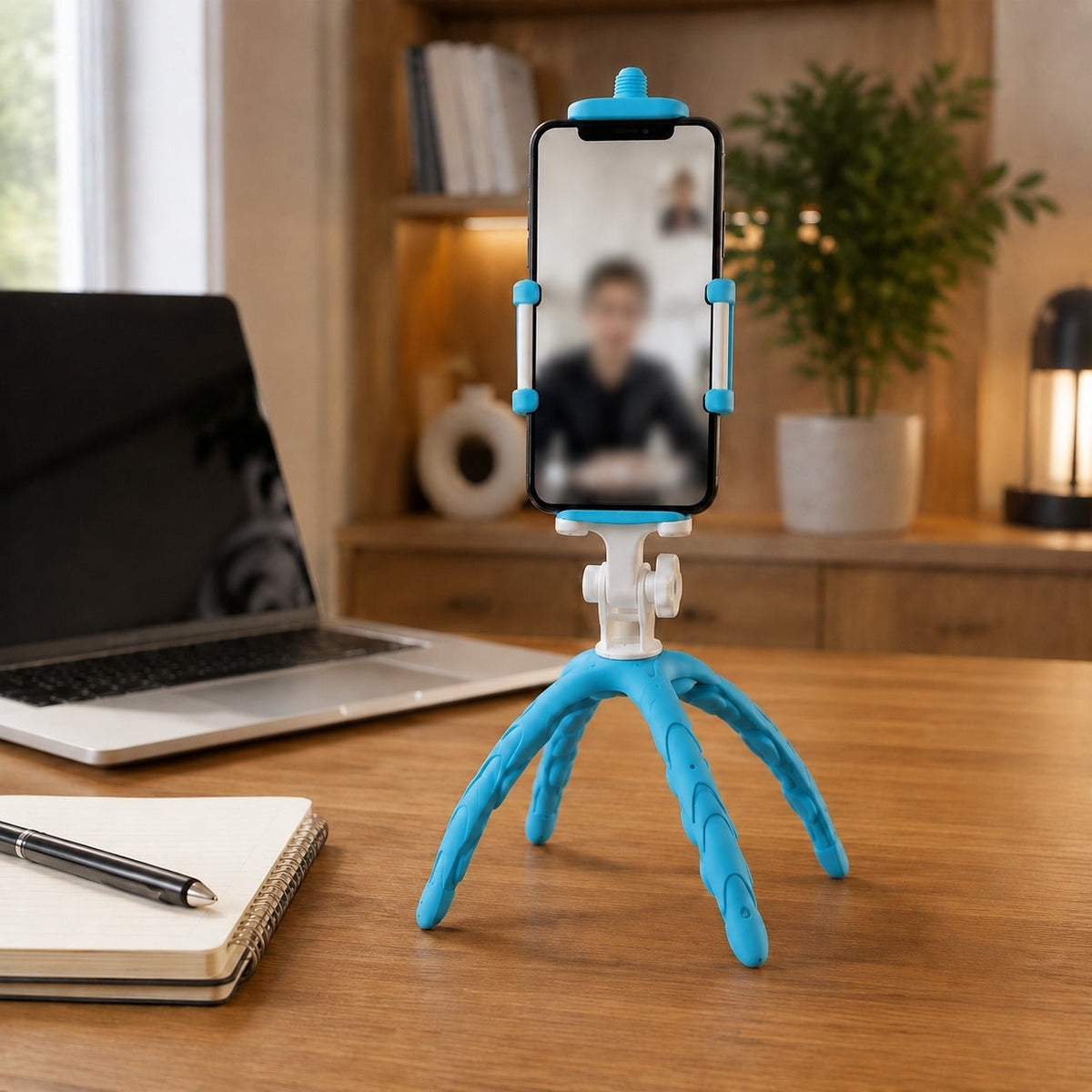 A blue and white flexible tripod holding a phone displaying a video call on a wooden desk with a laptop and notebook.