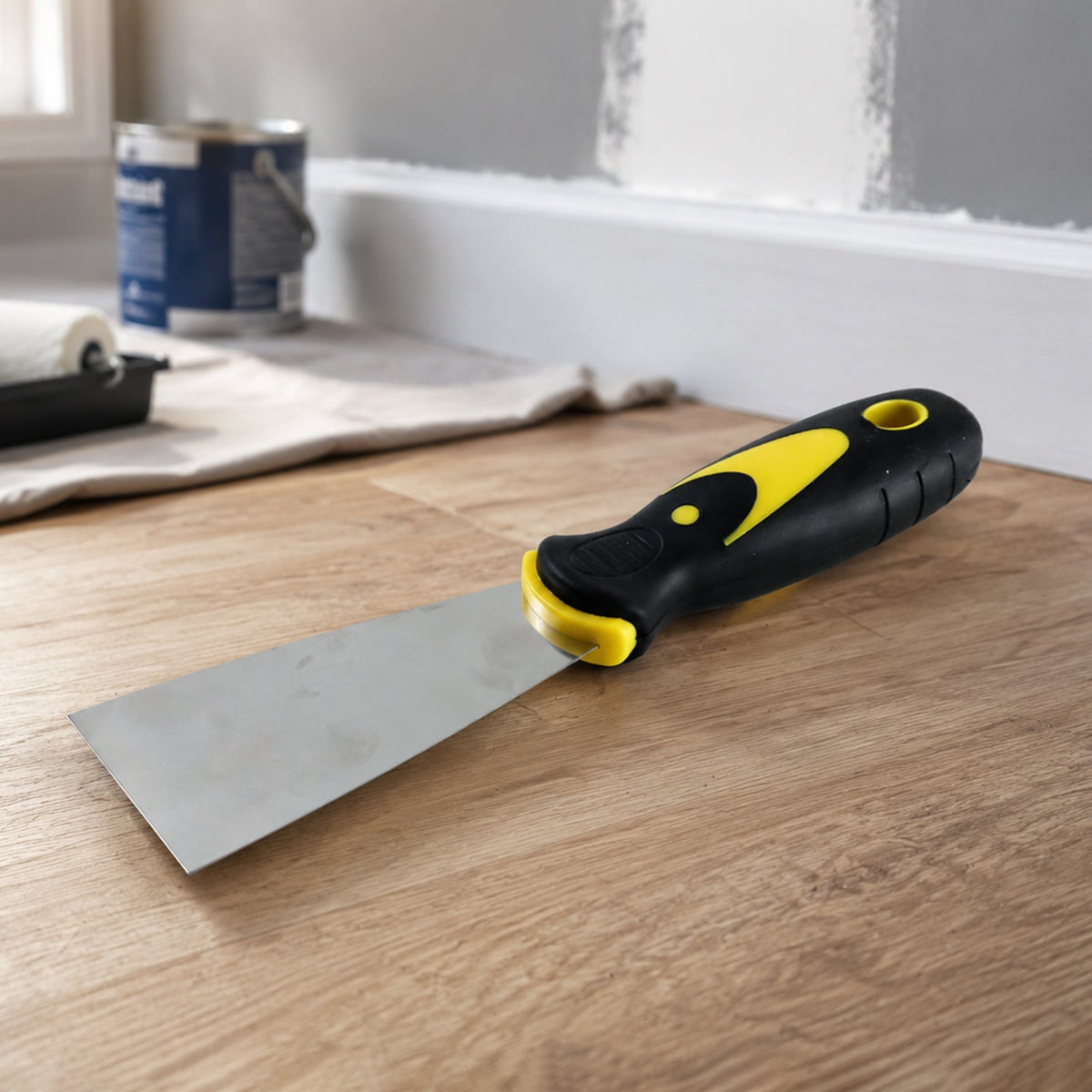 A putty knife with a black and yellow handle rests on a light wood floor next to a paint roller and can.