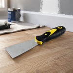 A putty knife with a black and yellow handle rests on a light wood floor next to a paint roller and can.