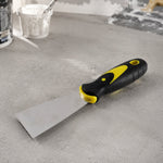 A putty knife with a black and yellow handle lies on a light gray concrete surface, with paint supplies in the background.
