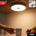A hand reaches for a mug under a bright, round LED light mounted beneath a wooden cabinet, illuminating a fruit bowl