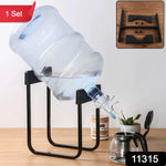 A large clear water bottle sits tilted on a black metal stand, dispensing water into a glass teapot, with a small inset